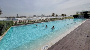 Ein Swimmingpool mit darin badenden Personen, umgeben von Liegestühlen und Sonnenschirmen, mit einer Stadtkulisse im Hintergrund.