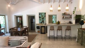 Un'ampia zona giorno con divano, tavolo da pranzo e sgabelli da bar. Si intravede in secondo piano una cucina con frigorifero e un quadro.