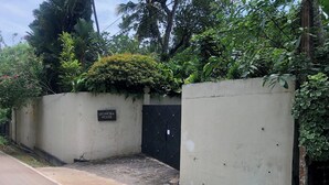 Un cancello chiuso dà accesso a una proprietà con l'insegna "LEONORA HOUSE", caratterizzata da un muro in cemento, un cancello nero e una vegetazione rigogliosa tutt'intorno.