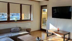 Ein modernes Wohnzimmer mit einer Couch, einem Holztisch, einem an der Wand befestigten Fernseher und einem großen Fenster mit Jalousien.