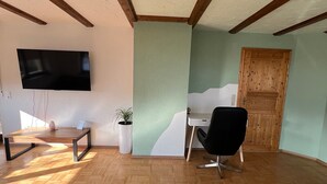 Ein Zimmer mit Holzboden, einem flachbildfernseher an der Wand, einem weißen Schreibtisch, einem schwarzen Bürostuhl und einer Holztür.