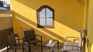 Eine Terrasse mit Steinböden, Holzboden und gelben Wänden. Zwei Stühle, ein Tisch und ein Blumentopf sind zu sehen.