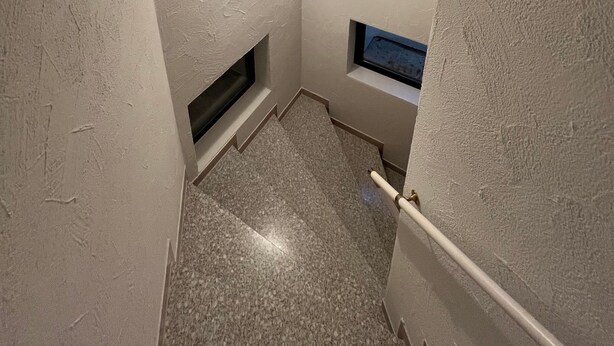 Eine schmale Treppe mit strukturierter Wand, einem kleinen Fenster und einer Lampe.