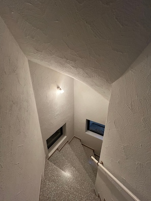 Un étroit escalier avec des murs à texture, une petite fenêtre et un luminaire.
