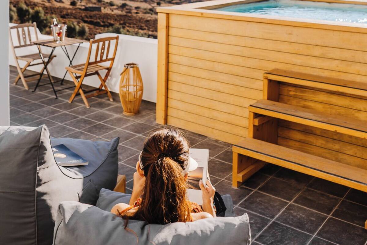 A person relaxing on a grey sofa with a view of a wooden hot tub and a pool, overlooking a coastal landscape.