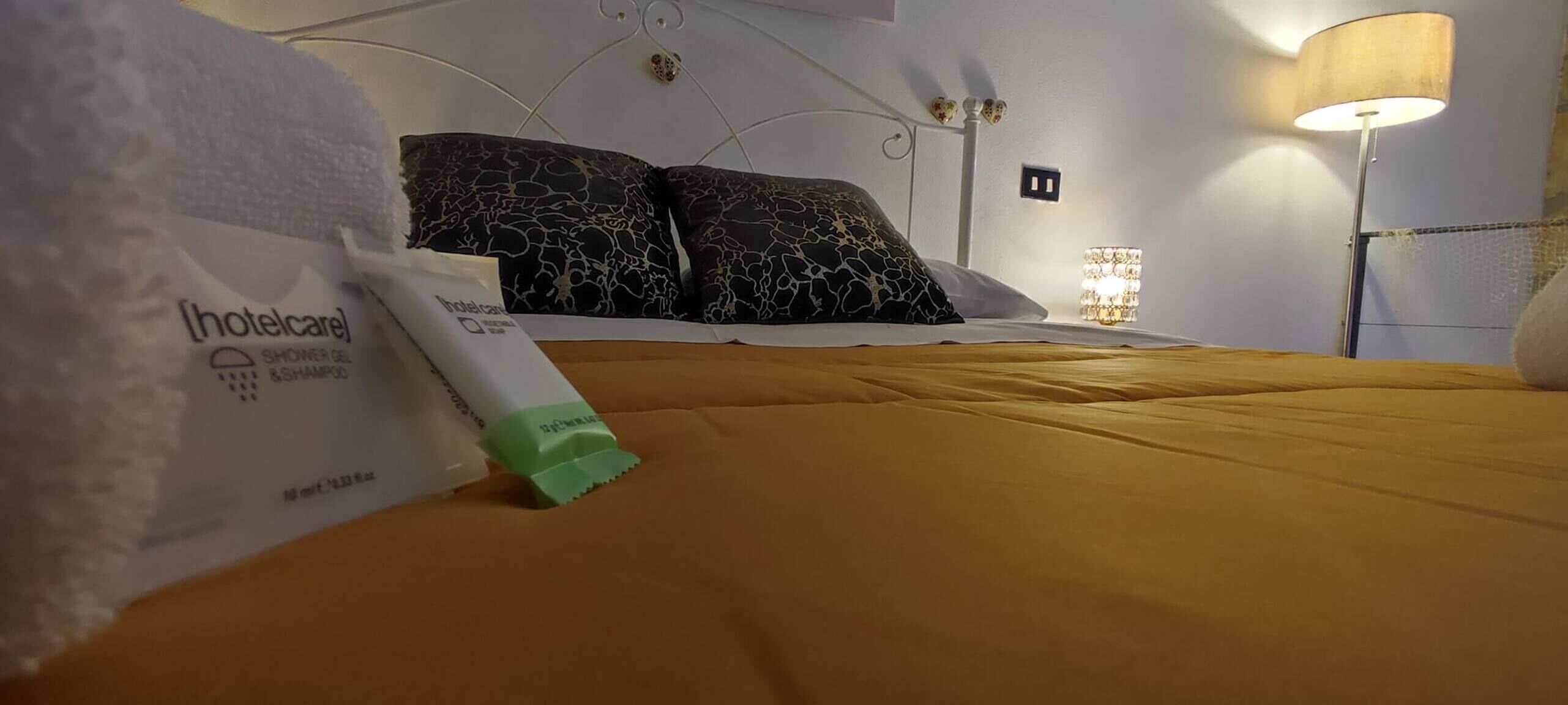 Camera d'albergo con un letto, cuscini, una lampada da comodino e un kit di cortesia dell'hotel sul letto.