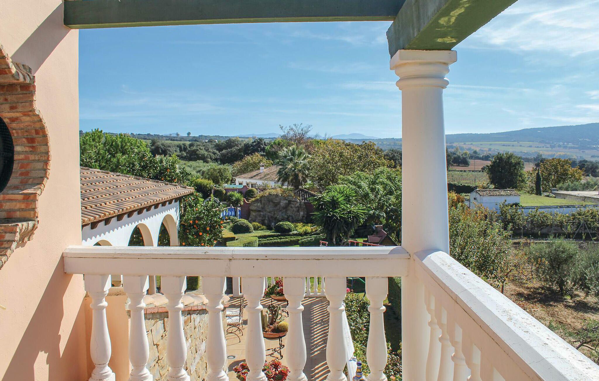 A balcony with a view of a landscaped garden, a building with a tiled roof, and a distant landscape with trees and hills.