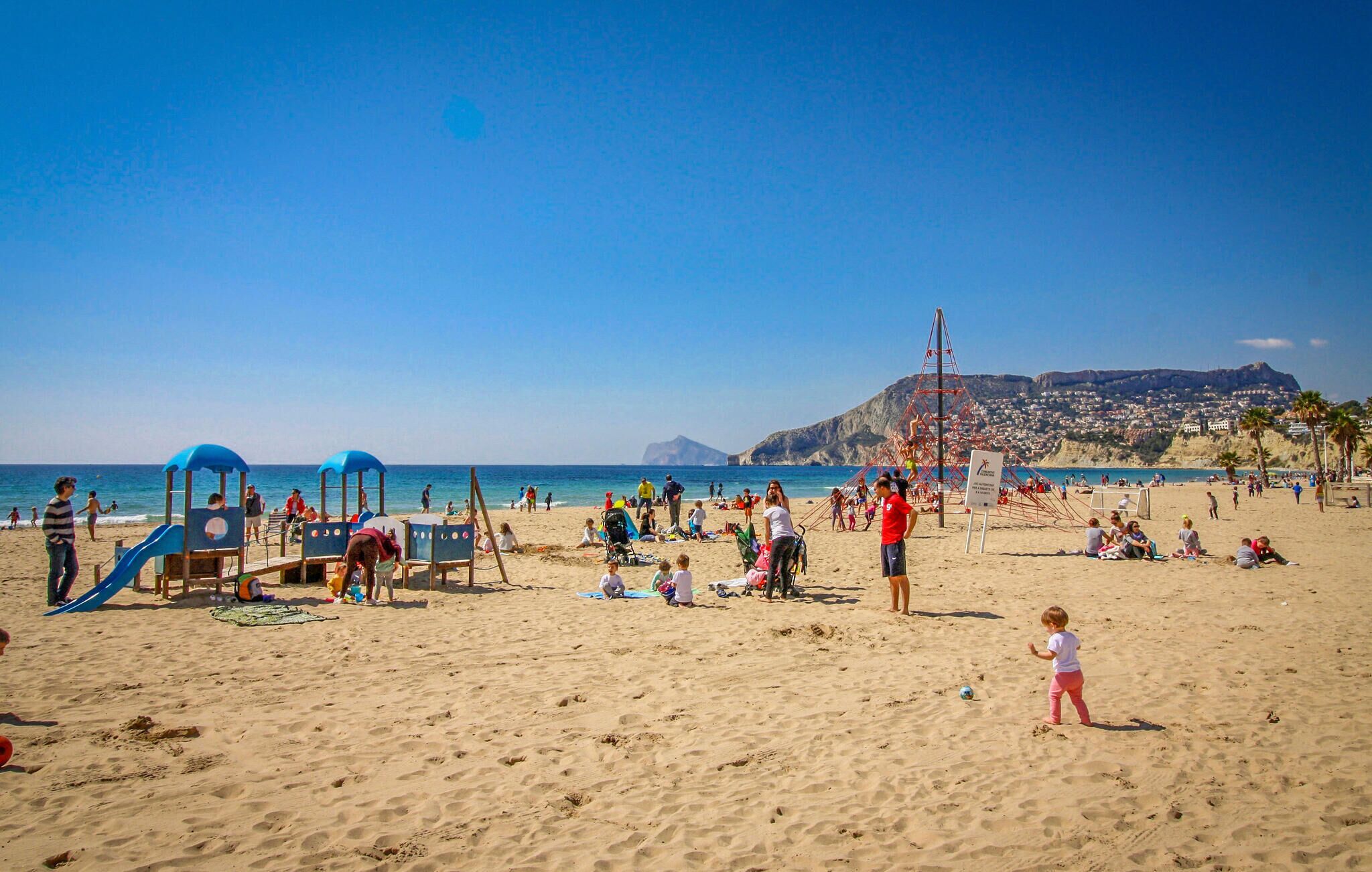 A beach with a playground, people relaxing, and a view of a town in the background.