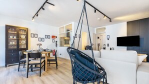 A modern living room with a white sofa, a hanging chair, a dining area with a table and chairs, and a television mounted on the wall. - 3-2-1meins Röntgen Remscheid – Modern Apartment for Up to 3 Guests (Remscheid)