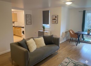 Een moderne woonkamer met een grijze bank, een eettafel en een zichtbare keuken door een open doorgang.