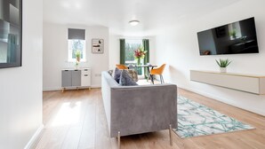 Una sala de estar moderna con un sofá gris, un televisor de pantalla plana, una zona de comedor con mesa y sillas, y una ventana con cortinas.