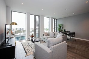 Un salon moderne avec un canapé gris, une table basse et un téléviseur. On y trouve également un coin repas avec une table et des chaises. De grandes fenêtres offrent une vue sur la ville.