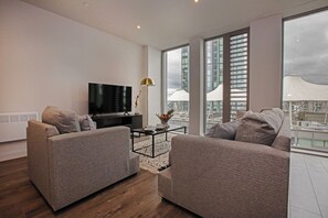 Un salon moderne avec deux canapés gris, un téléviseur noir, une table basse et un lampadaire. De grandes fenêtres offrent une vue sur le paysage urbain.