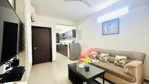 A living room with a sofa, coffee table, and a TV. There is a kitchen in the background with cabinets and a refrigerator. - BluO 1BHK Jaipur Terrace Garden Lift (Jaipur)