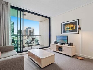 A modern living room with a sofa, a coffee table, a TV on a stand, and a floor lamp. There is a large window with a view of buildings. - High-Floor 1BR City Escape with Balcony Views (Brisbane)