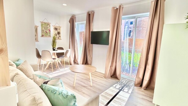 Un salon moderne comprenant un canapé, une table à manger avec des chaises, un téléviseur fixé au mur et de grandes fenêtres avec des rideaux.