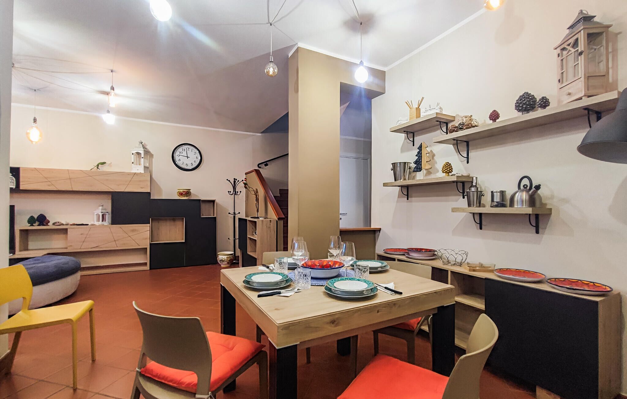 Un coin repas avec une table en bois, dressée pour quatre personnes, entourée de chaises dotées de coussins orange. Une horloge est fixée au mur au-dessus de la table.