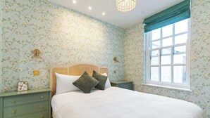 Een slaapkamer met bloemenbehang, een houten hoofdbord, een nachtkastje in het groen en een raam met uitzicht.