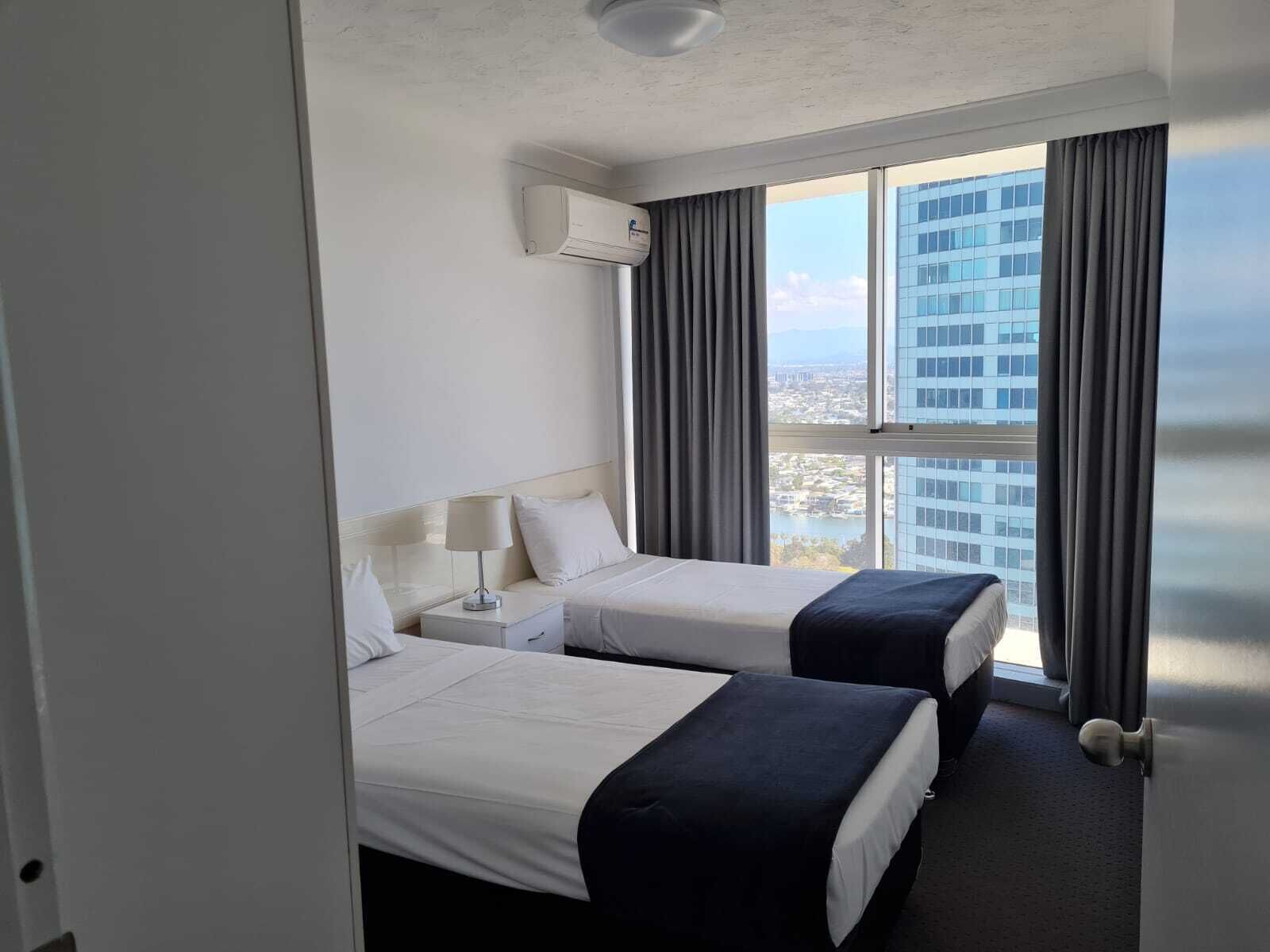 A hotel room with two beds, a nightstand, a lamp, and a view of a cityscape through large windows.