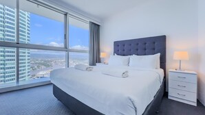 A hotel room with a large bed, a nightstand, and a view of a cityscape through large windows. - Level 38 Ocean-View Apartment, Surfers Paradise (Surfers Paradise)