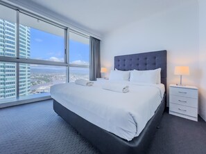 Ein Hotelzimmer mit einem großen Bett, einem Nachttisch und Blick auf eine Stadtlandschaft durch große Fenster.