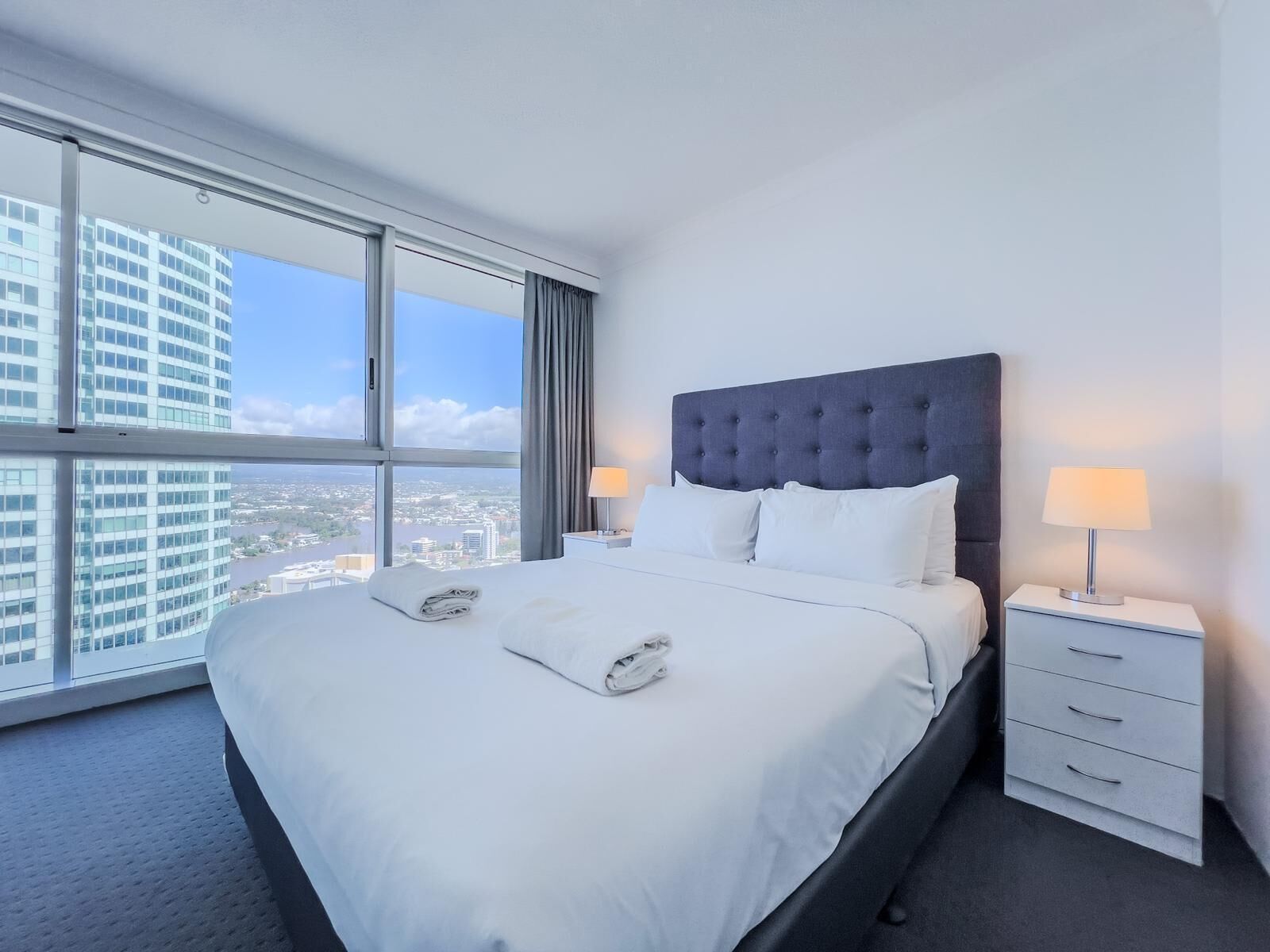 Una habitación de hotel moderna con una cama grande, dos mesitas de noche con lámparas y vistas a un paisaje urbano a través de amplios ventanales.