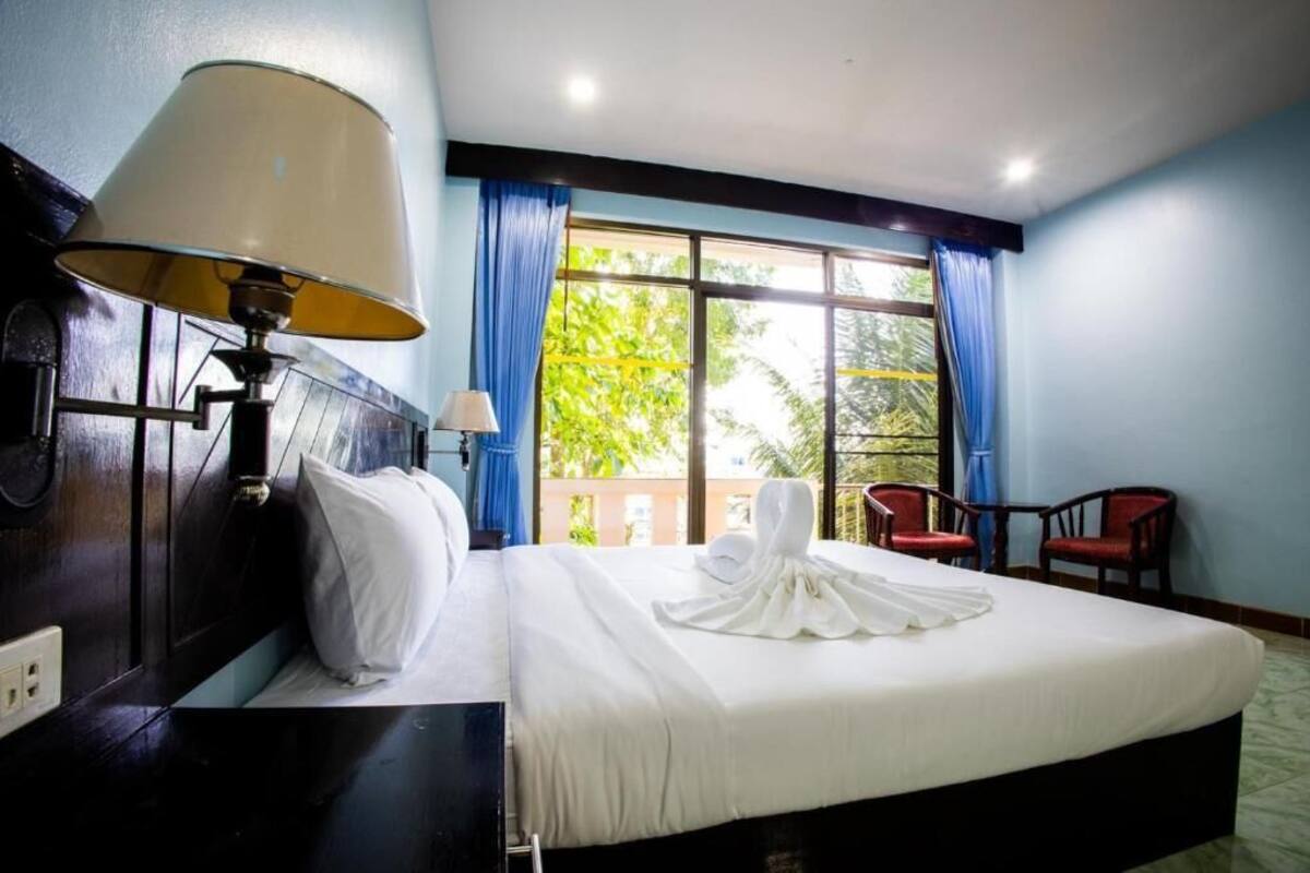 Une chambre d’hôtel avec un grand lit, une table de chevet avec une lampe, une fenêtre avec des rideaux bleus et une vue sur la verdure.