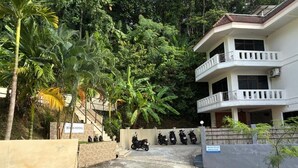 A building with a gated entrance, surrounded by lush greenery and parked motorcycles. - Riviera Guesthouse Patong (Patong)