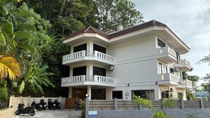 A two-story building with balconies, a parking area with motorcycles, and lush greenery around. - Riviera Guesthouse Patong (Patong)