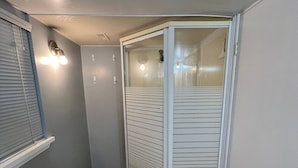 Un couloir étroit avec une cabine de douche en verre, une fenêtre avec des stores et un luminaire fixé au mur.