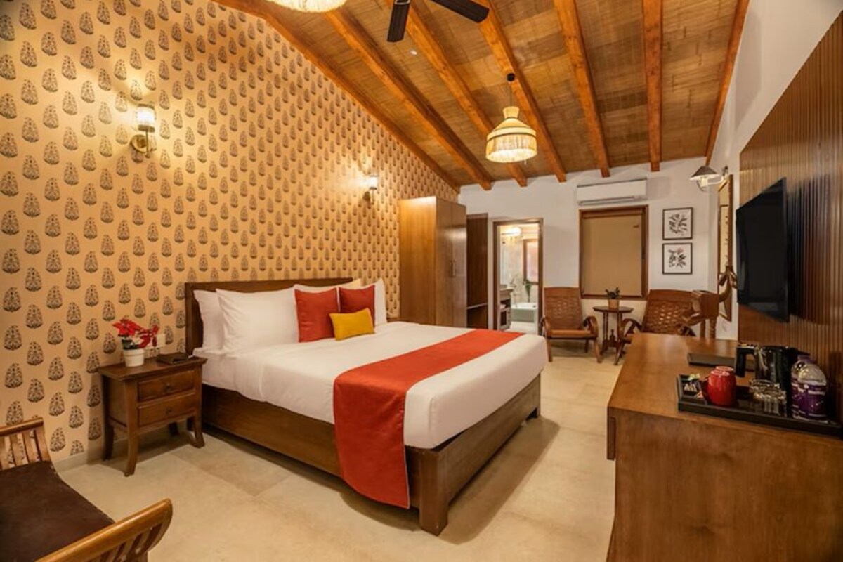 A hotel room with a wooden ceiling, a bed with a red blanket, a wooden nightstand, a desk with a chair, and a TV on the wall.