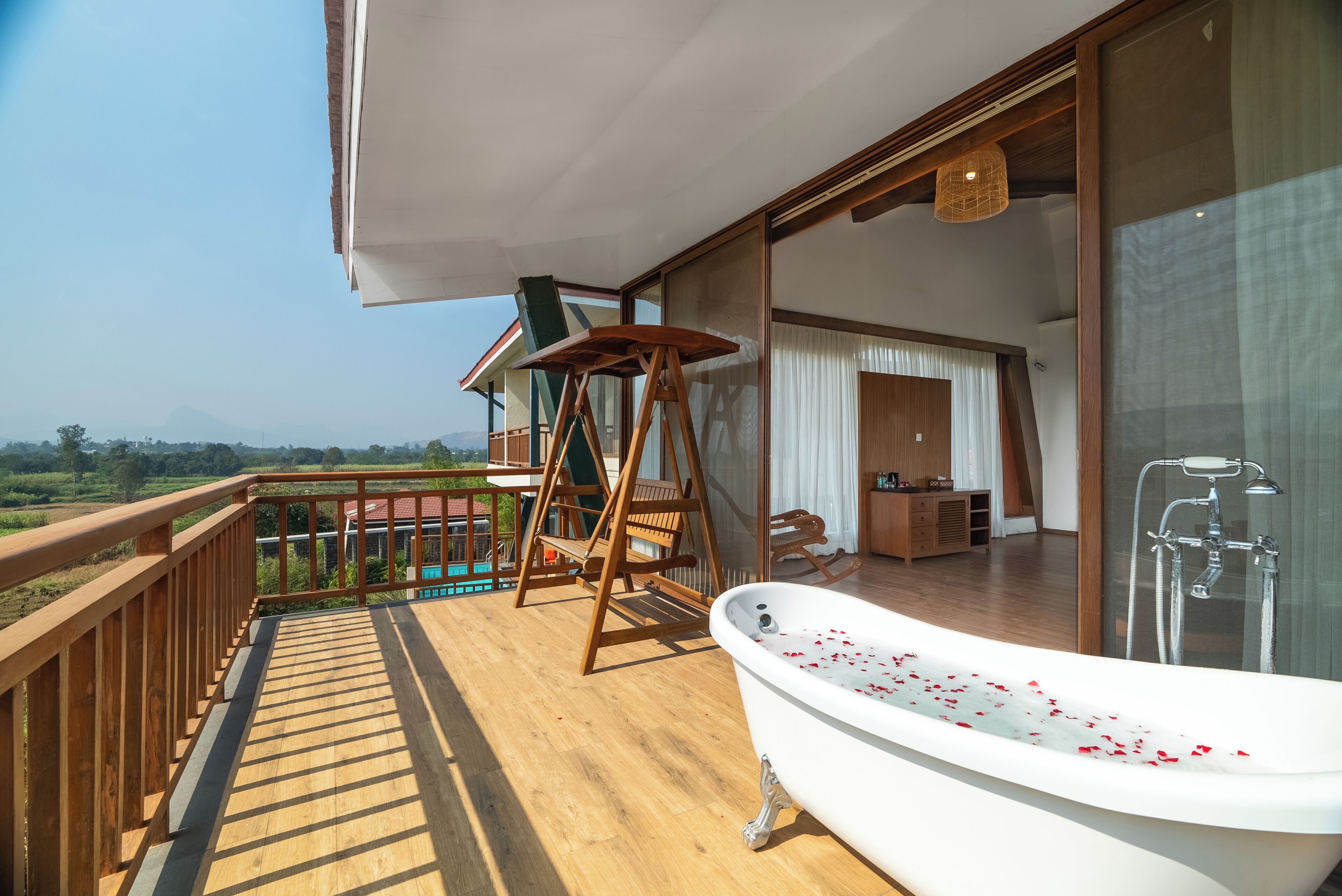 Un balcon avec un plancher en bois, une baignoire blanche parsemée de pétales rouges et un fauteuil suspendu.