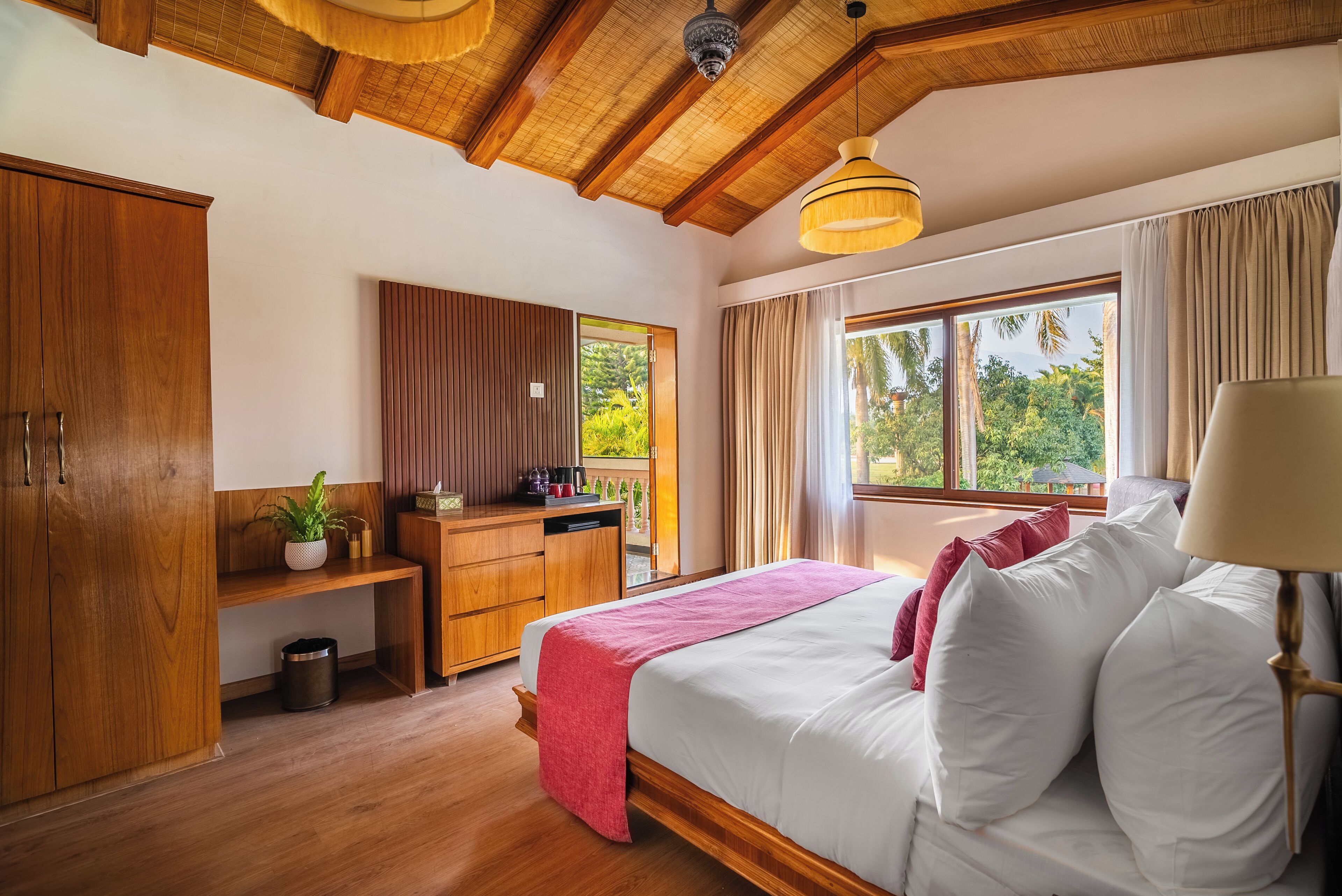 A well-lit bedroom with a wooden ceiling, a bed with white linens, a bedside table, a desk with a chair, and a window with curtains.