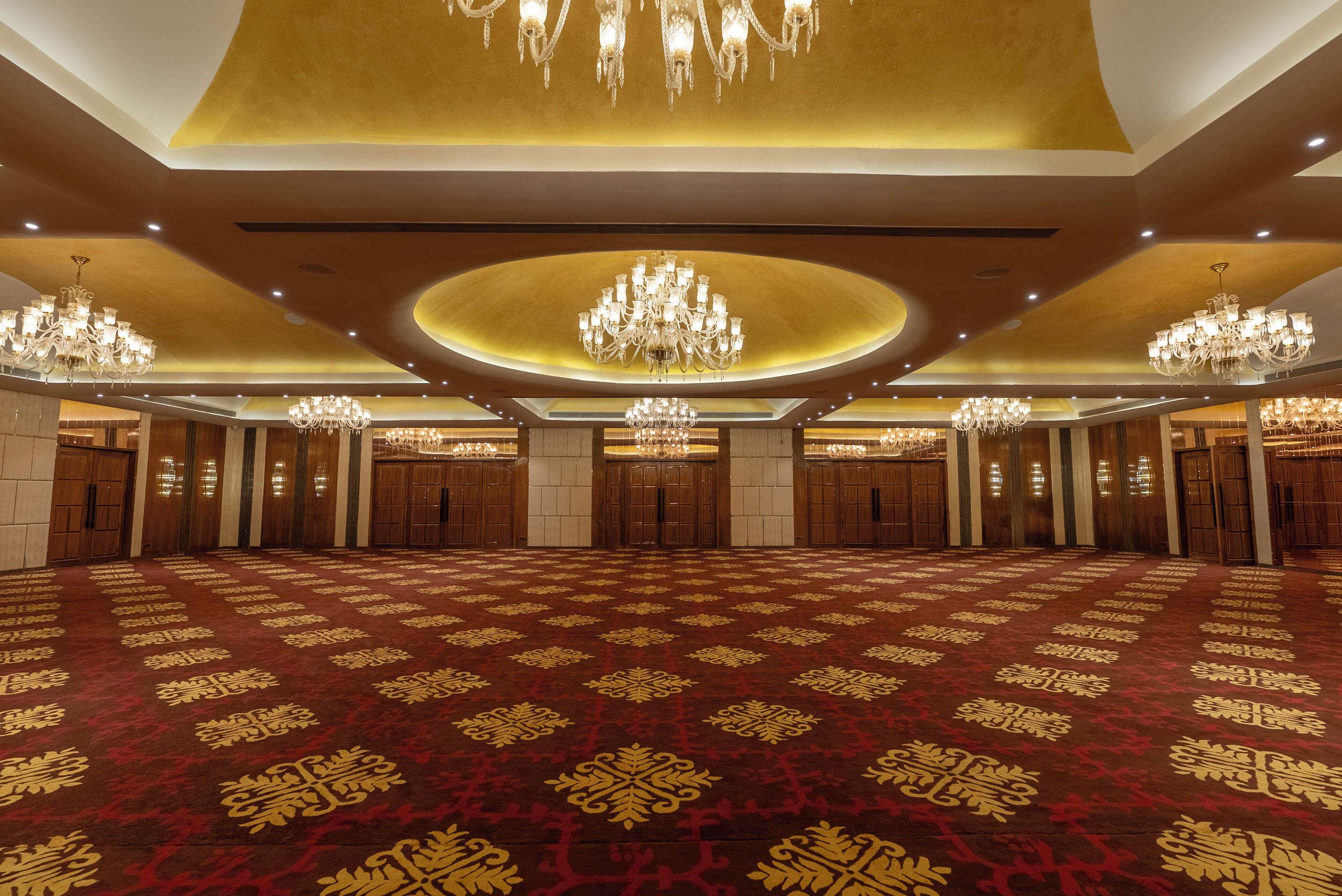 A spacious hall with a patterned carpet, multiple chandeliers, and wooden doors.