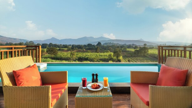 Un espace aménagé au bord de la piscine, avec des meubles en osier, une table sur laquelle sont disposés une assiette de nourriture et des boissons, et une vue sur un paysage montagneux.