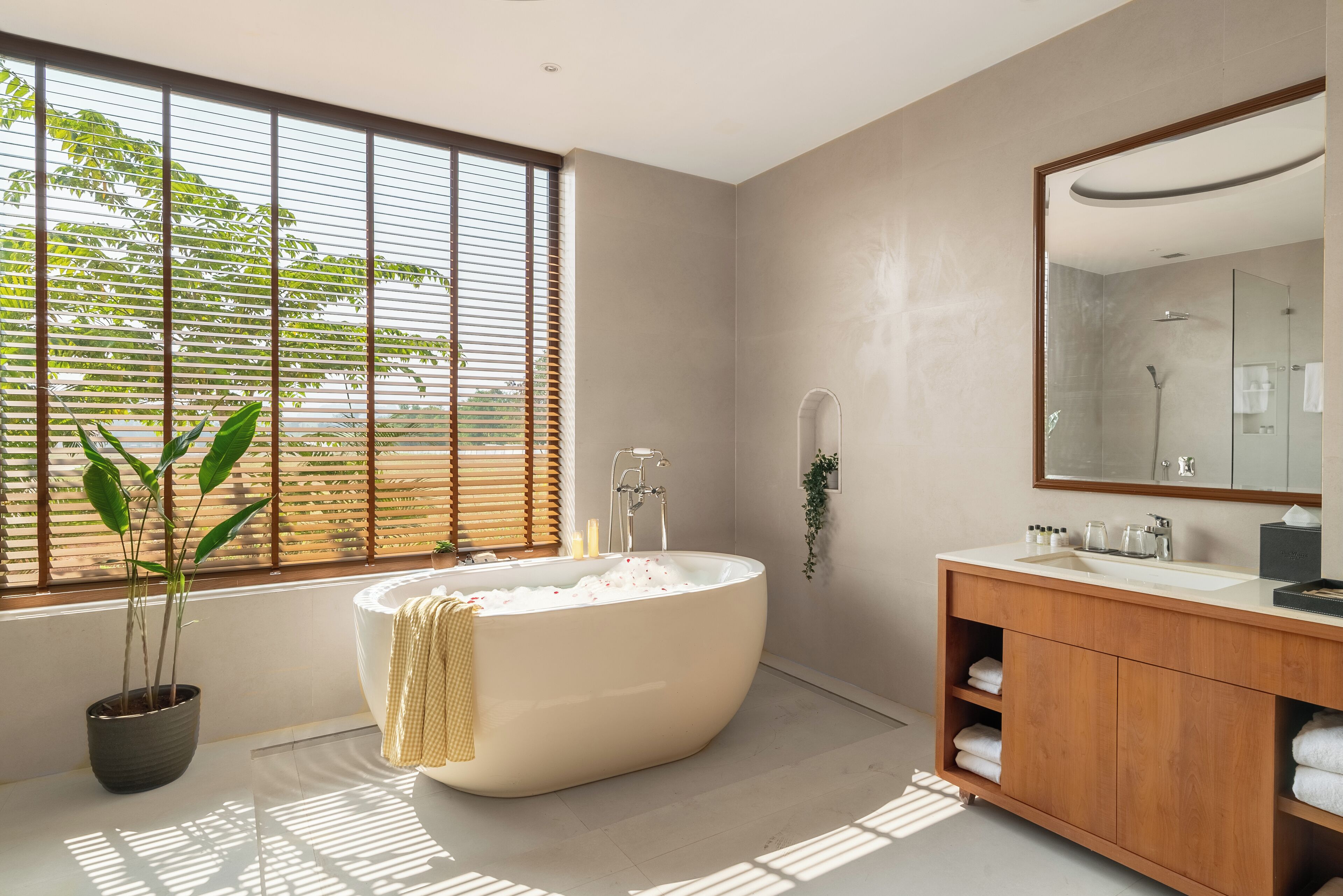 A modern bathroom with a freestanding bathtub, a wooden vanity, a large mirror, and a window with blinds.