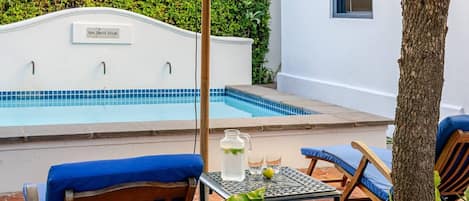 Un área junto a la piscina con una pared blanca, una piscina con azulejos azules y dos sillas con cojines azules.