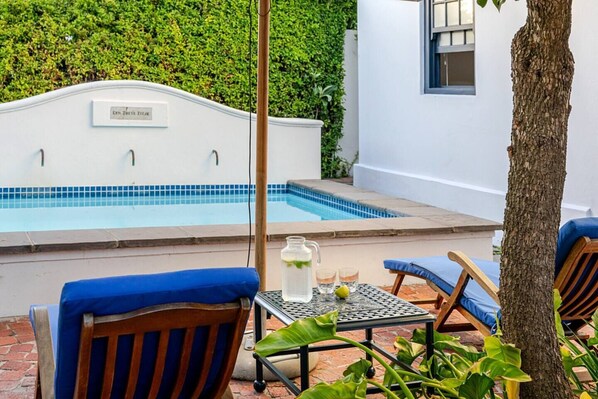 A poolside area with a white wall, a blue-tiled pool, and two blue cushioned chairs.