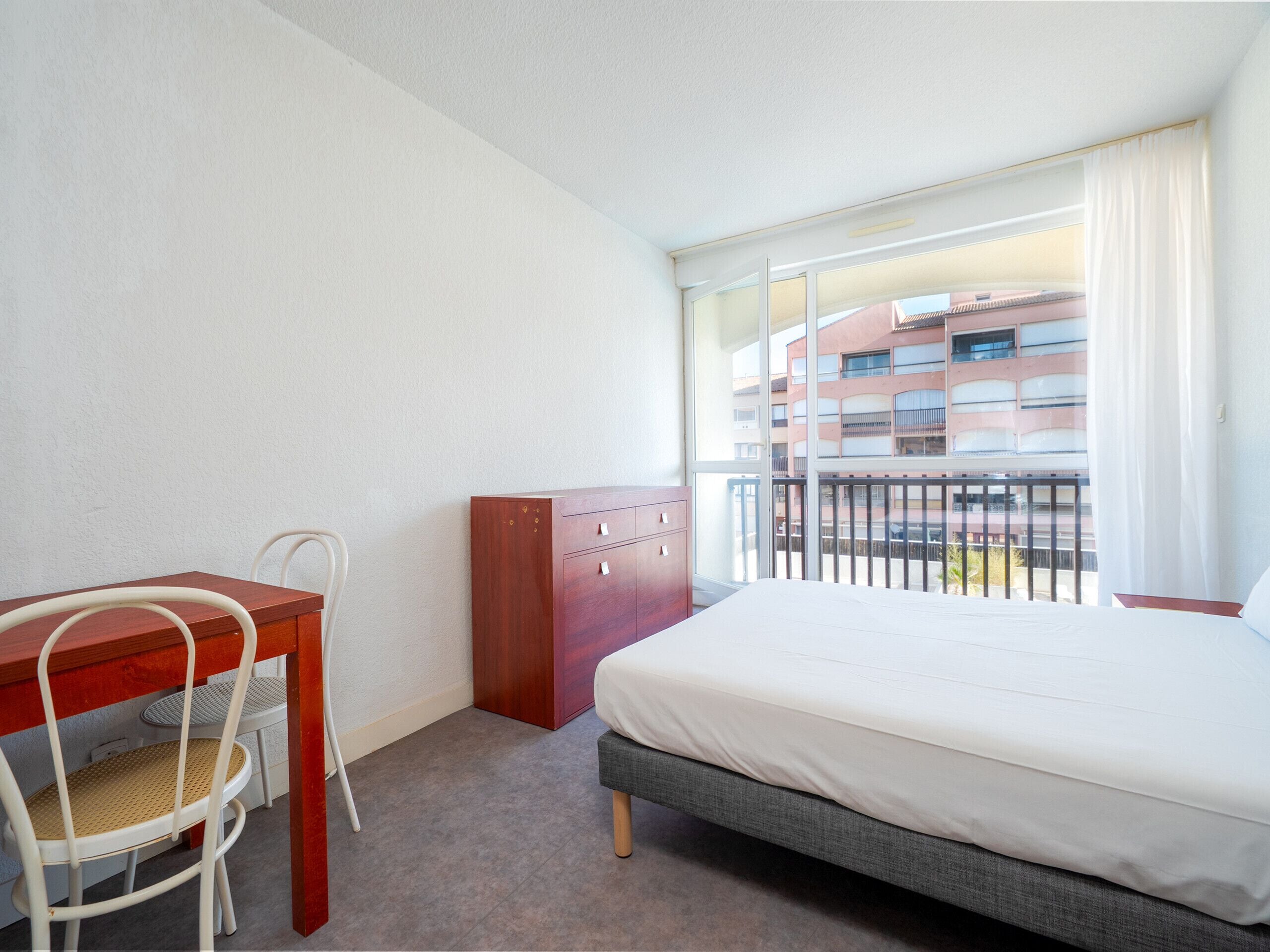 Ein Hotelzimmer mit einem Bett, einem Schreibtisch und einem Stuhl. Ein Fenster bietet Blick auf umliegende Gebäude.
