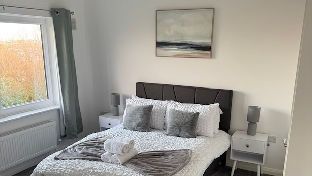 A neatly made bed with a white and gray quilt, a gray throw blanket, and a rolled towel. There is a gray lamp on a bedside table, a gray lamp on the other side, and a painting on the wall. - Rowe Terrace Workington (Workington)