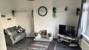A living room with a grey sofa, a coffee table with flowers, a TV on a stand, and a clock on the wall. - Rowe Terrace Workington (Workington)