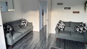 A modern living room with two grey sofas, each with geometric patterned cushions, and a wooden floor. There are wall-mounted signs with phrases like "this is us" and "family". - Rowe Terrace Workington (Workington)