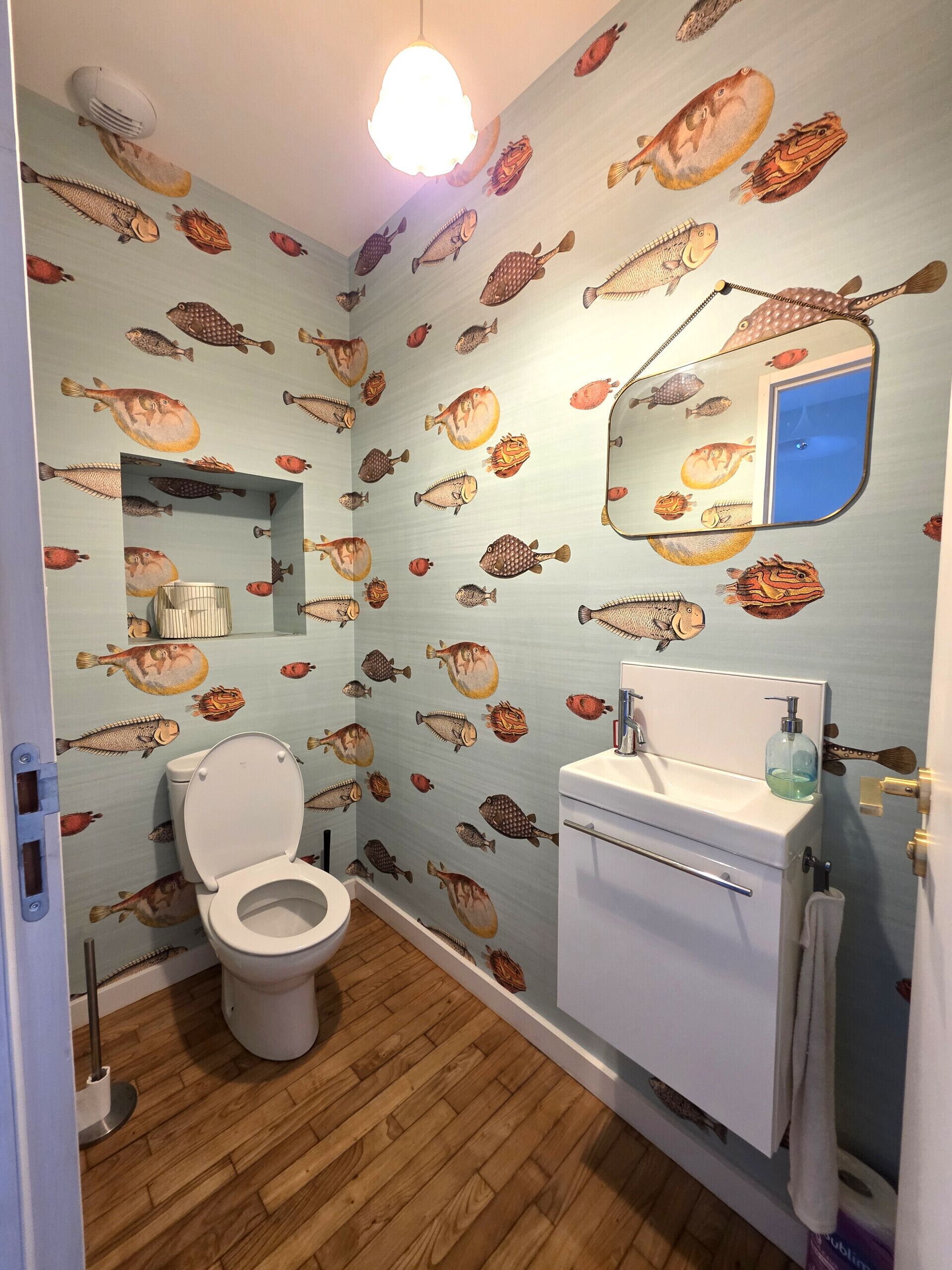 Une salle de bain comprenant des toilettes, un lavabo et un miroir. Les murs sont décorés de motifs représentant des poissons.