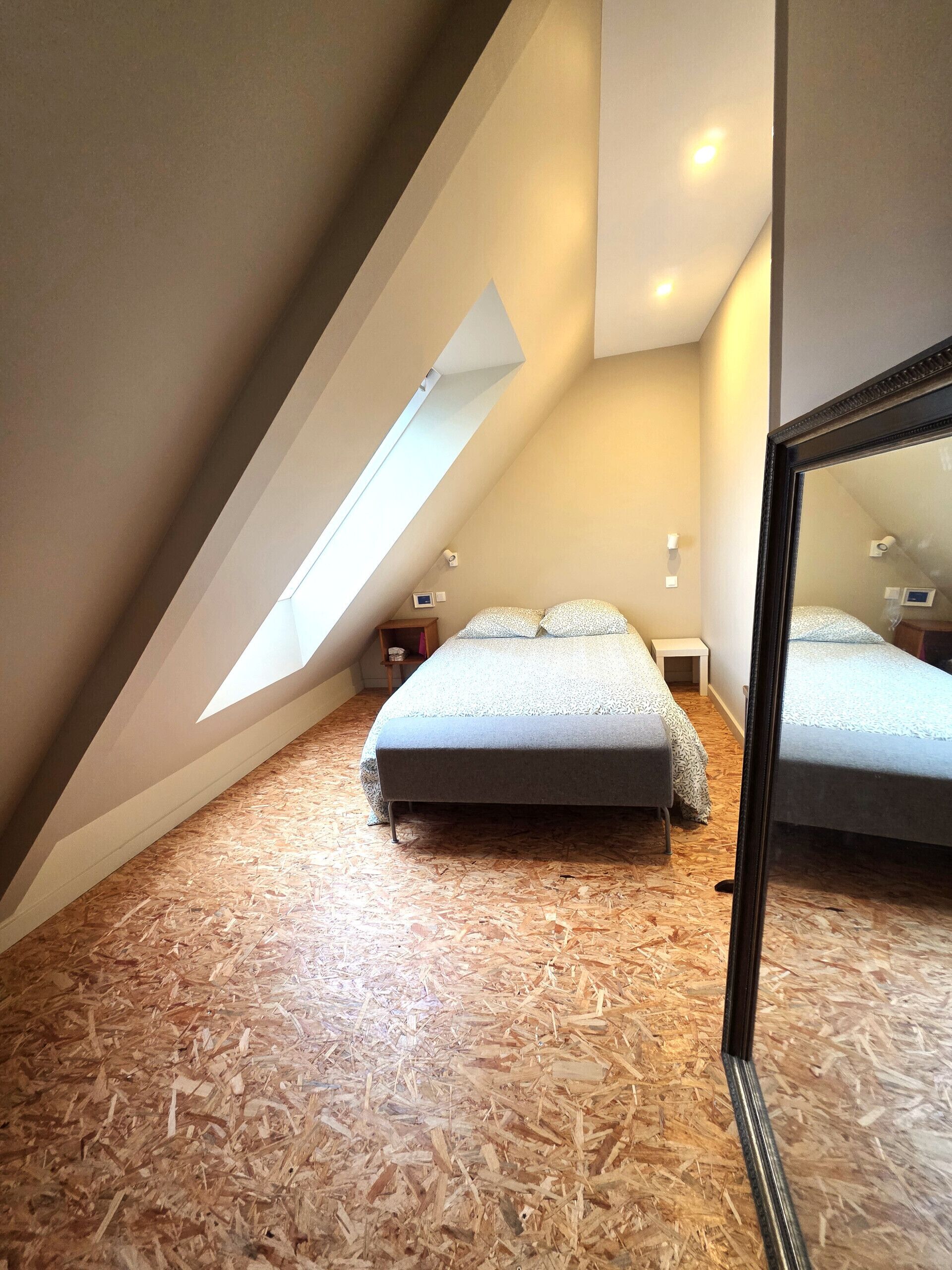 Une chambre avec un plafond en pente, un puits de lumière, un lit, une table de chevet et un miroir.