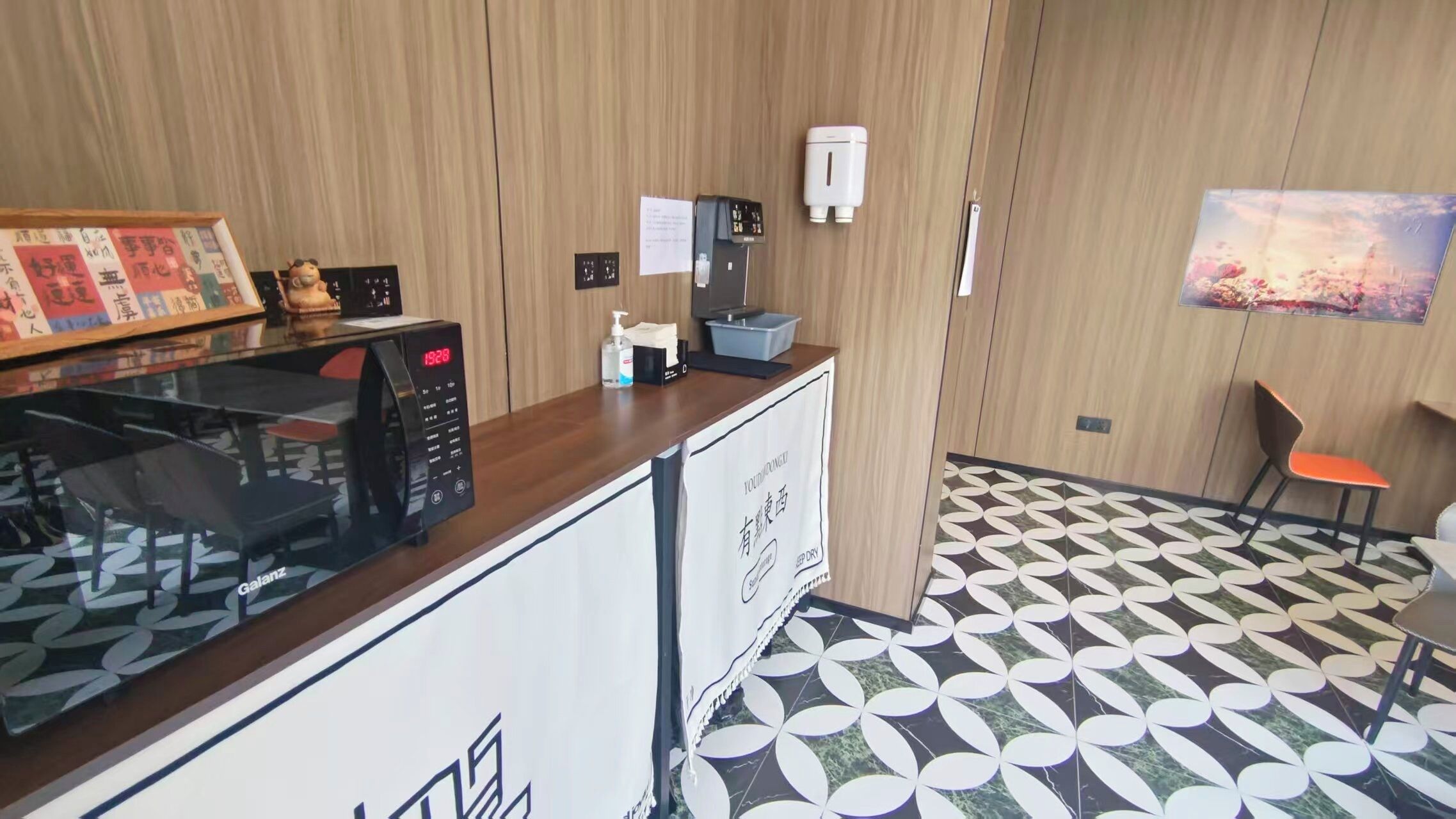 A hotel room with a wooden panel wall, a microwave, a small table with a chair, a hand sanitizer dispenser, and a painting on the wall.