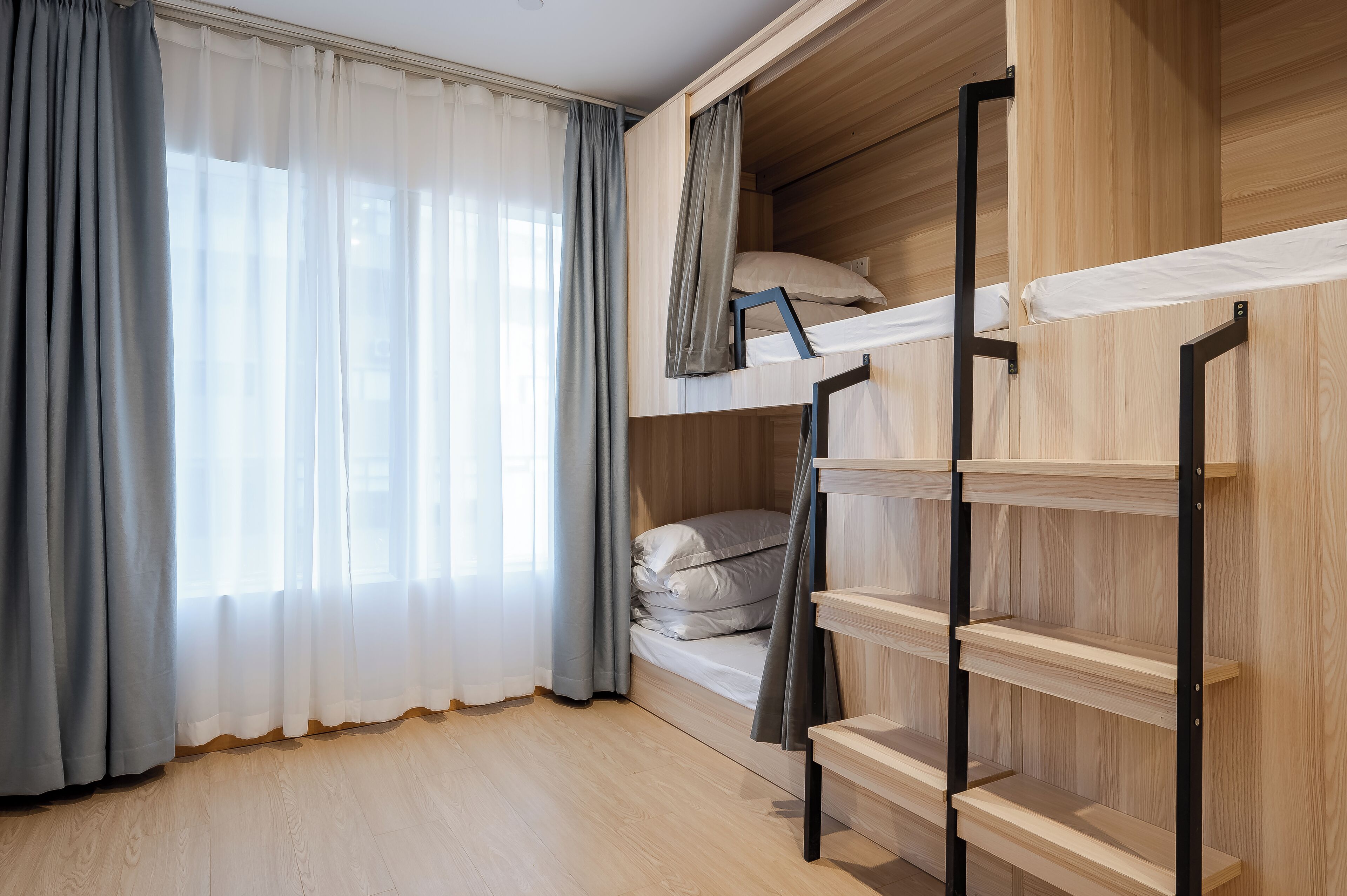 A bunk bed room with a wooden floor, a window with sheer curtains, and a built-in shelf unit.