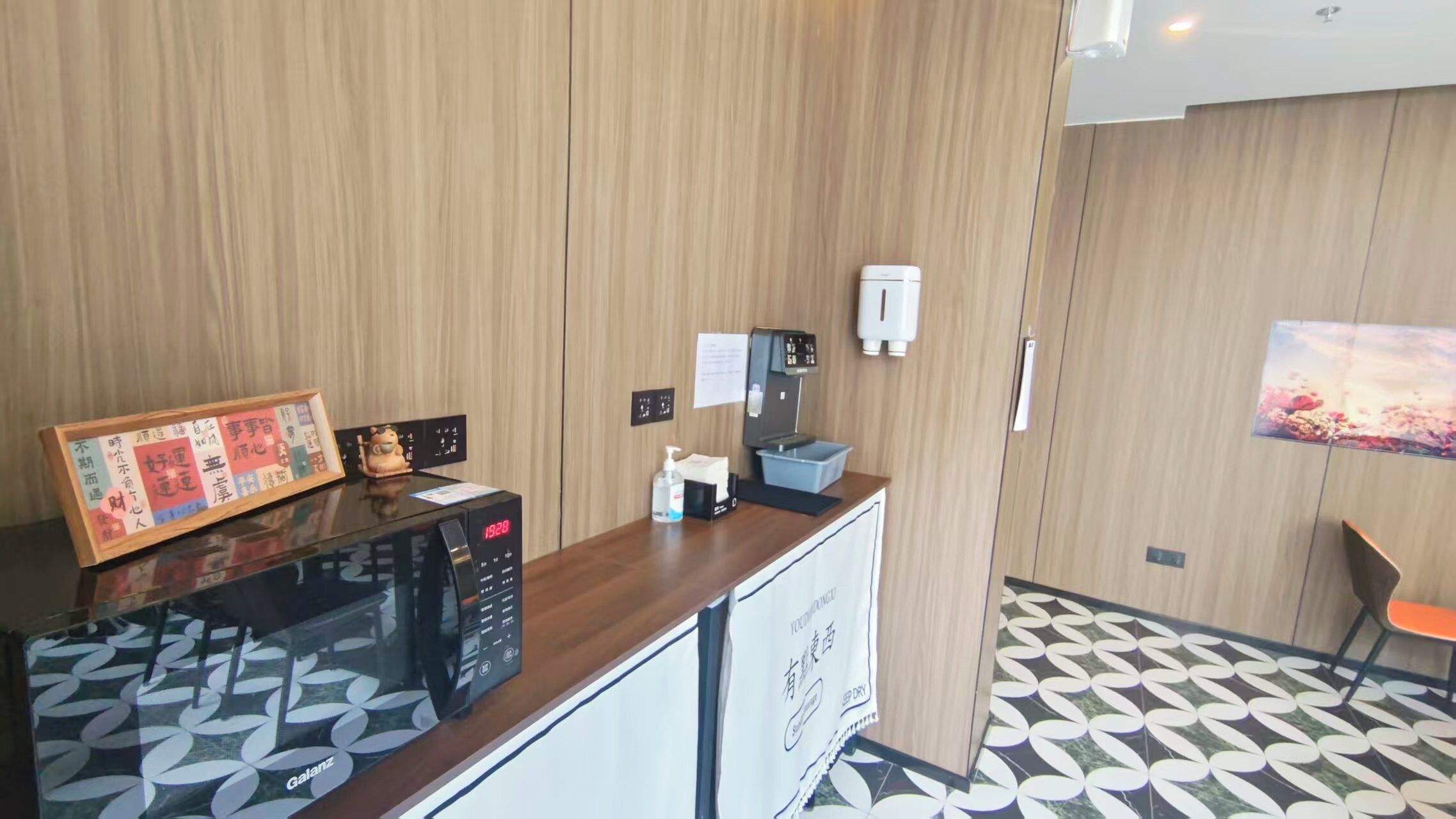 A hotel reception area with a wooden panel wall, a digital clock, a coffee machine, a small table with a framed picture, and a patterned floor.