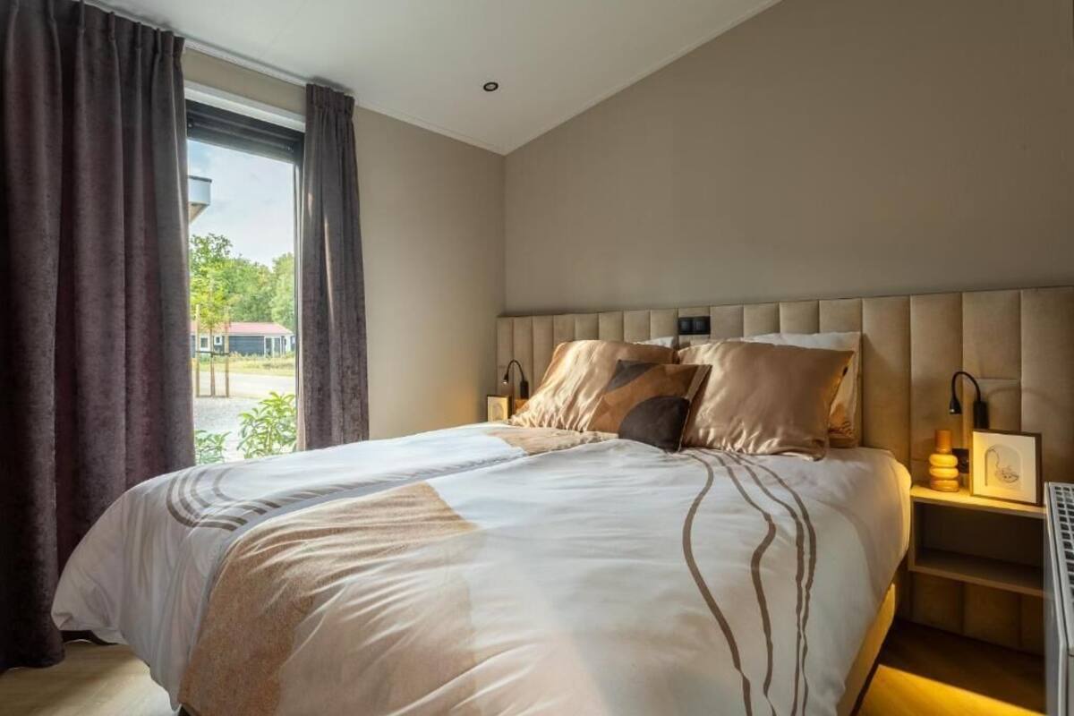 Een net opgemaakt bed met een beige hoofdbord, een nachtkastje met een lamp, en uitzicht op groen buiten.