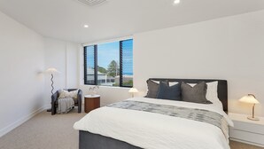 A bedroom with a large bed, two bedside tables, a chair, and a window with a view of the beach.