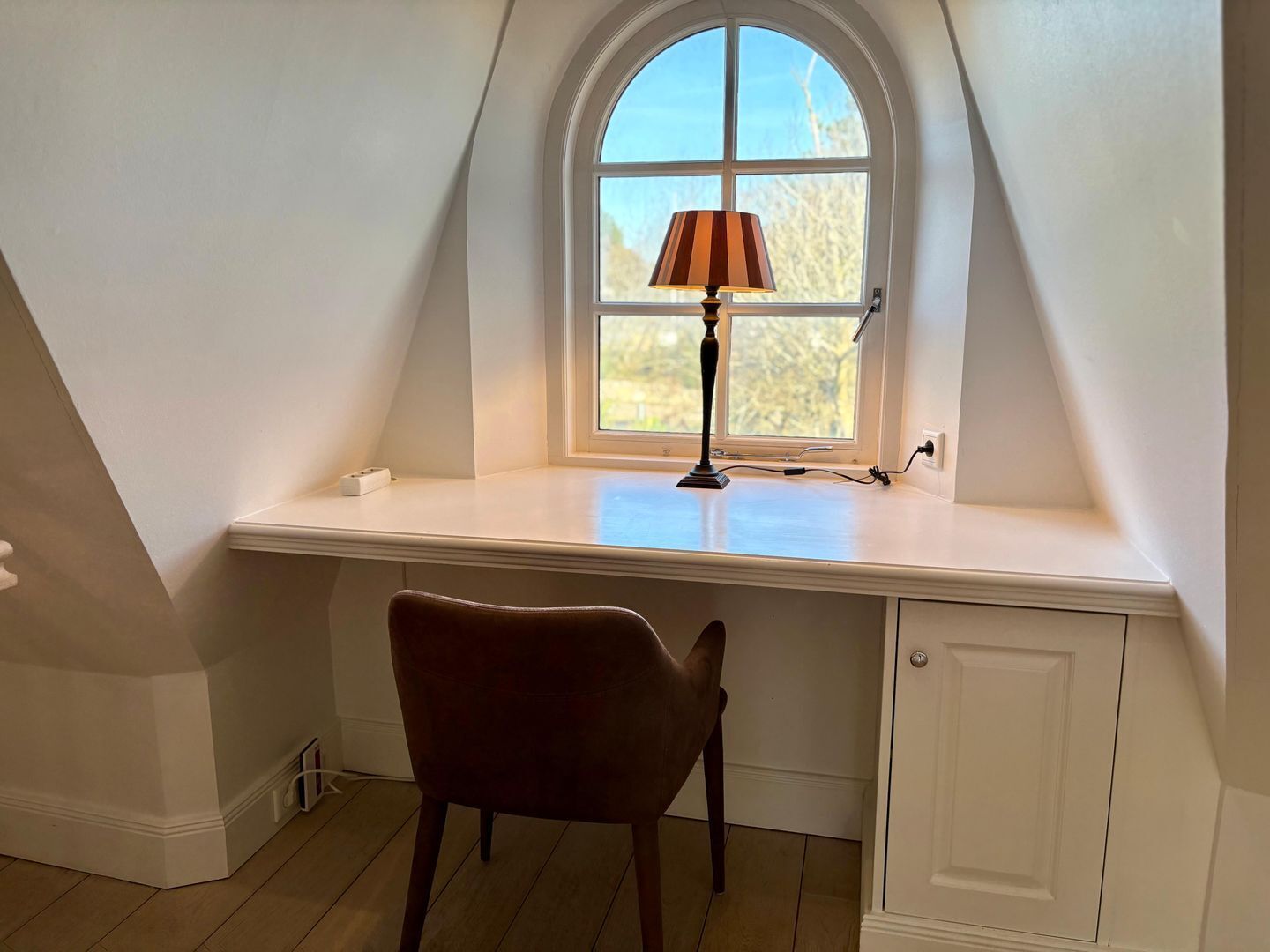 A room with a sloped ceiling, a desk with a lamp, a chair, and a door.